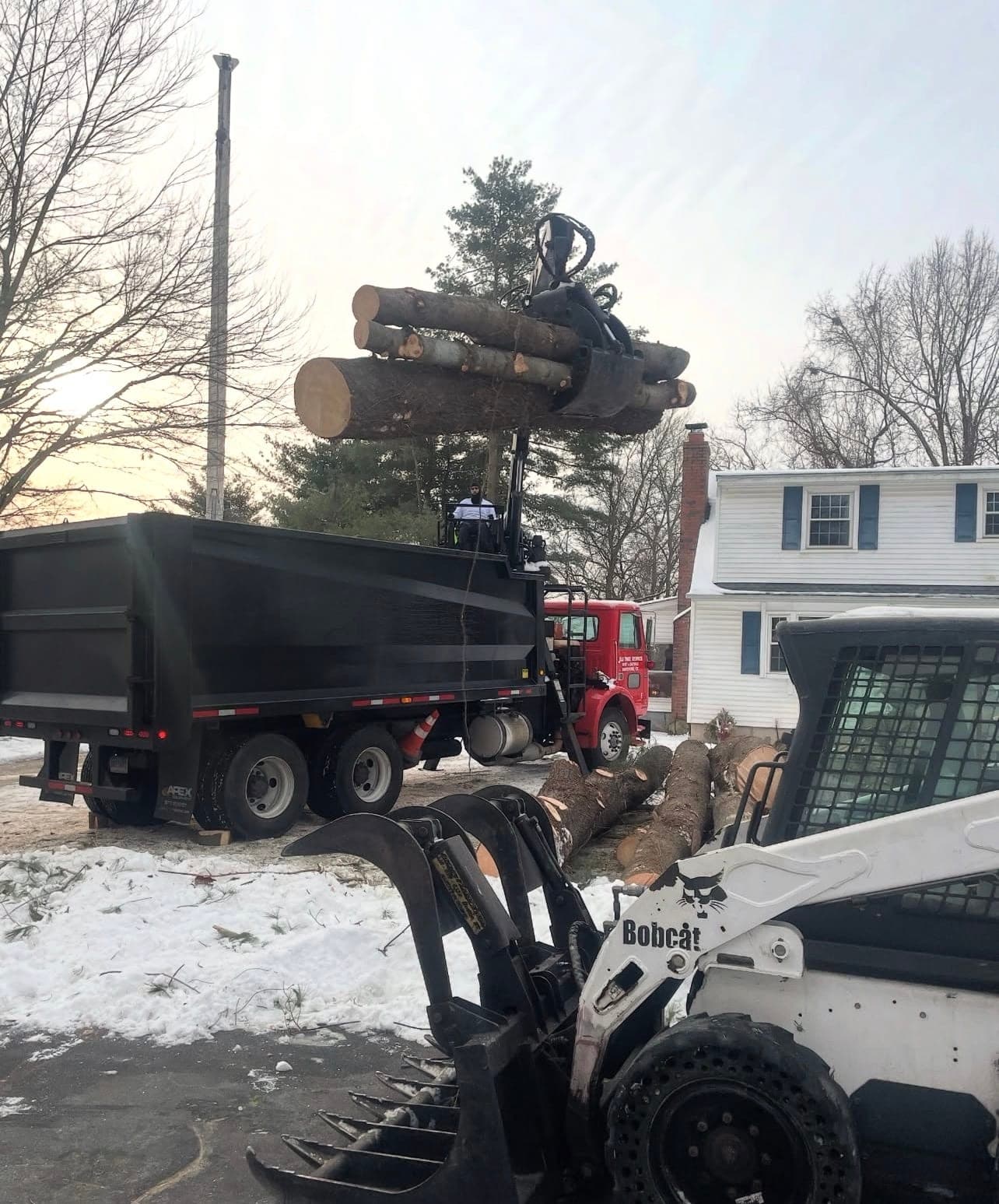 Skid steer with a grapple lifting logs into a dump truck in the snow