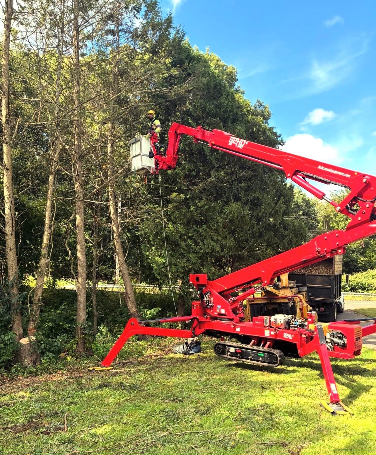 Climber in the bucket of a fully extended red spider lift at the edge of a wooded yard